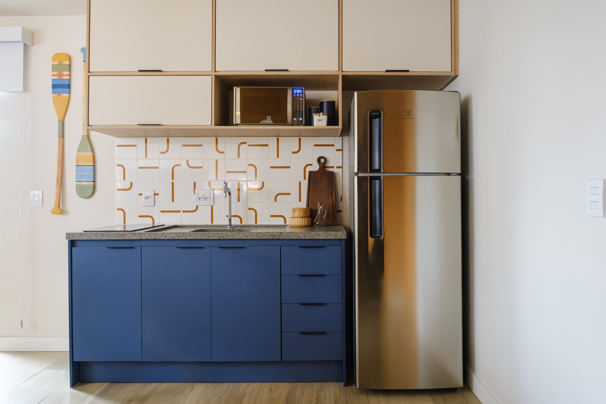 Compact kitchen with blue lower cabinets, white and terracotta geometric backsplash, microwave in the niche and stainless-steel fridge.