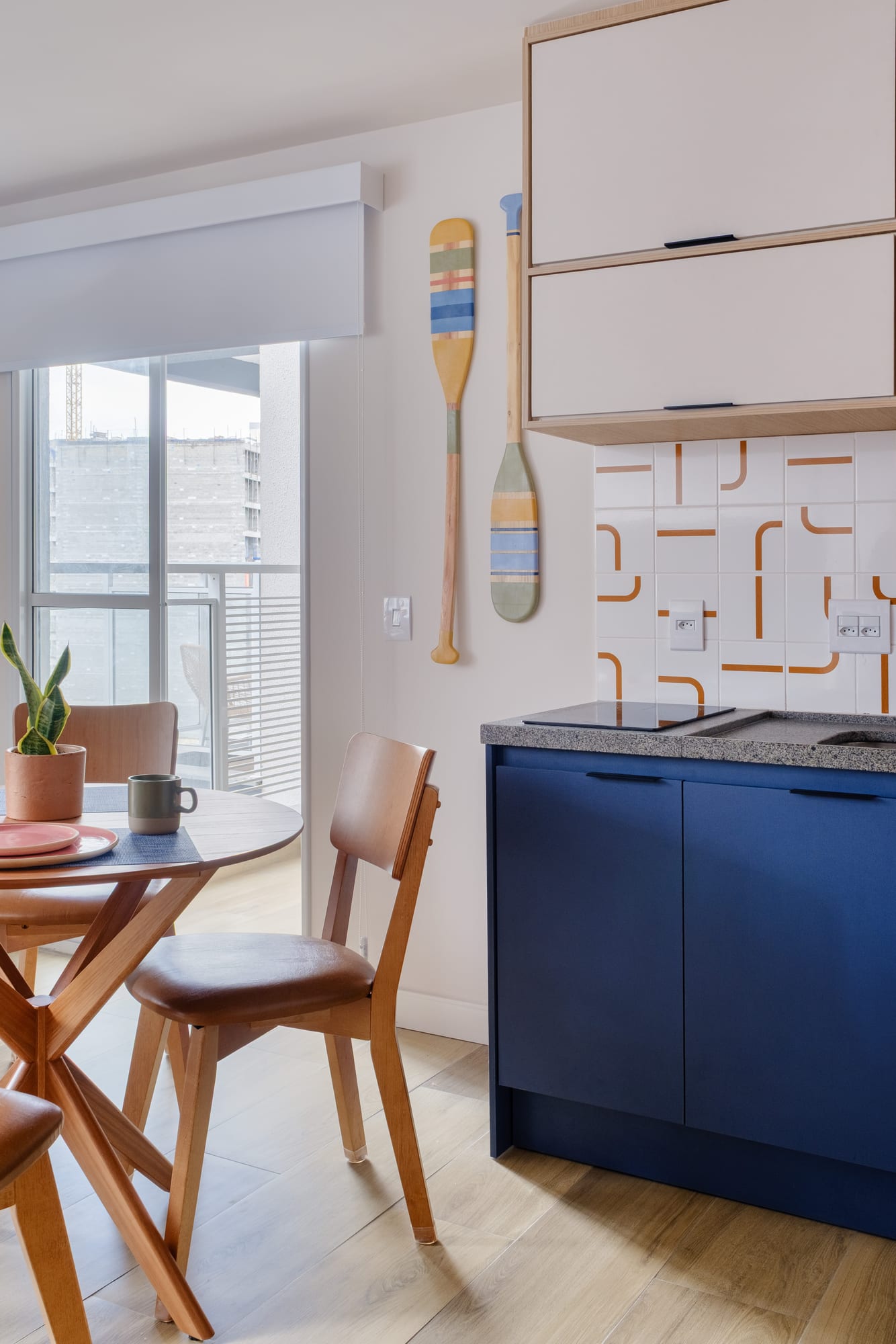Round wooden dining table with chairs beside the blue kitchen, decorative paddles on the wall and a glass door to the balcony.