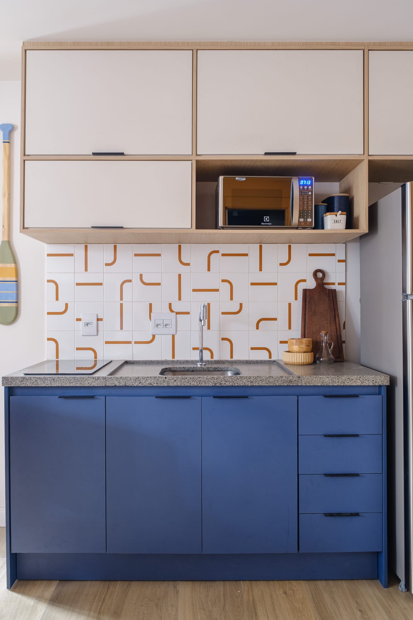 Front view of the kitchen with blue cabinetry, granite countertop, sink, cooktop and terracotta graphic tile.