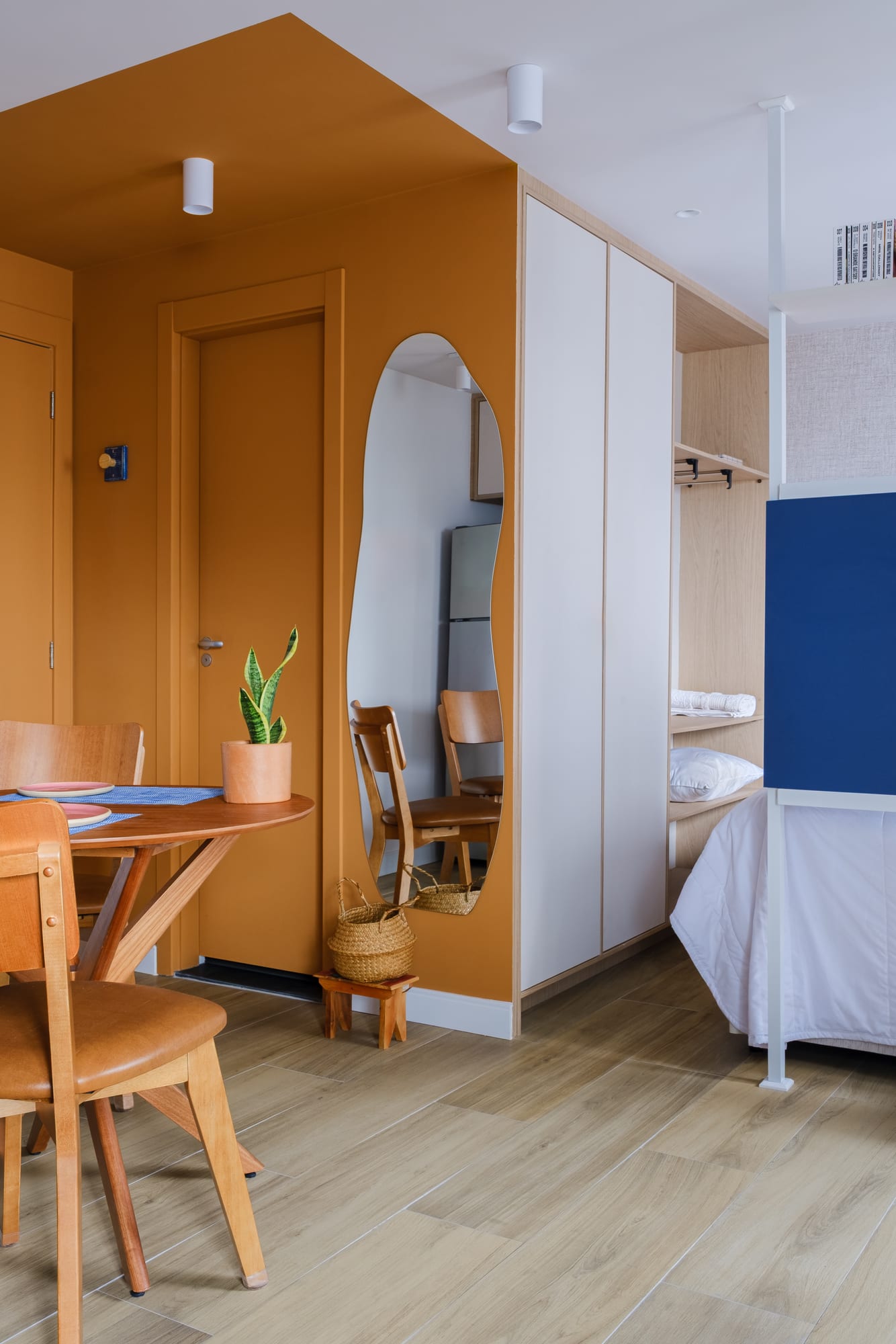 Mustard-toned entry hall with an organic mirror, wooden dining table and the studio bed in the background.