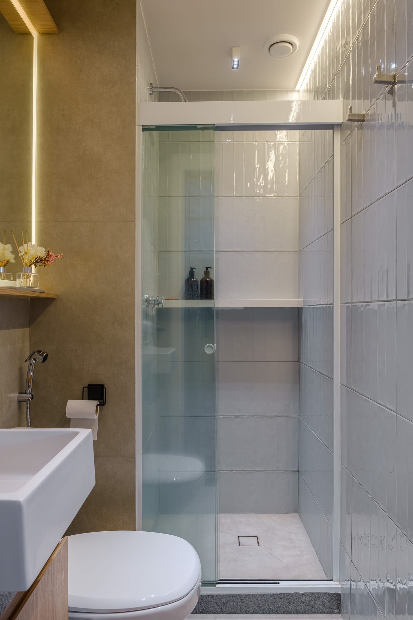 Bathroom shower with glossy light-toned tile, a niche with amenities and a white sink with a backlit mirror.