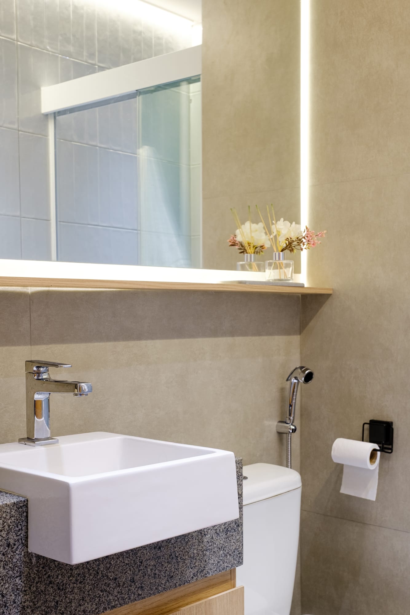 White sink on a granite countertop with a backlit mirror, toilet and toilet paper on a black holder.