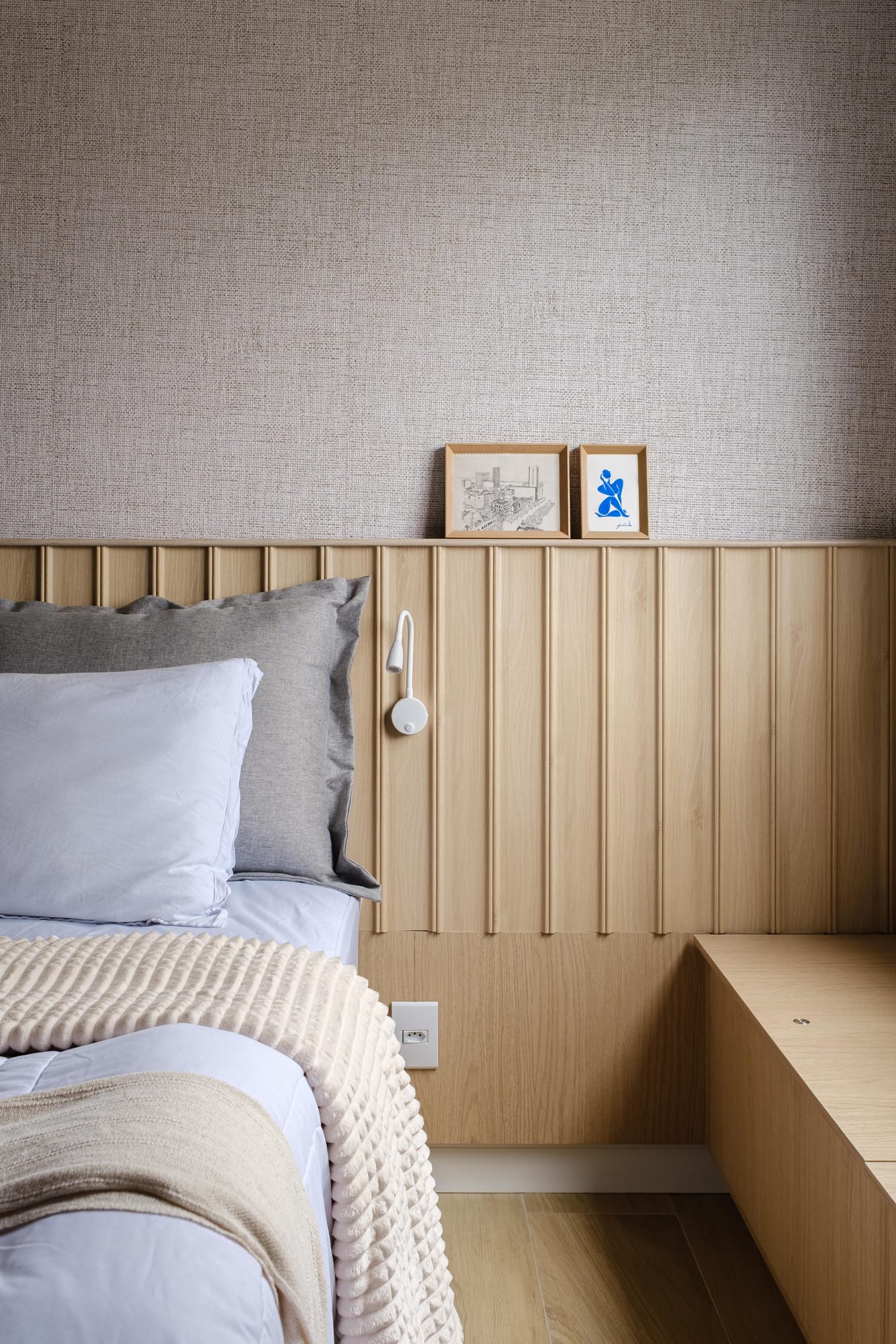 Bed detail with white linens and a textured throw, slatted wood headboard and framed prints resting on the shelf.