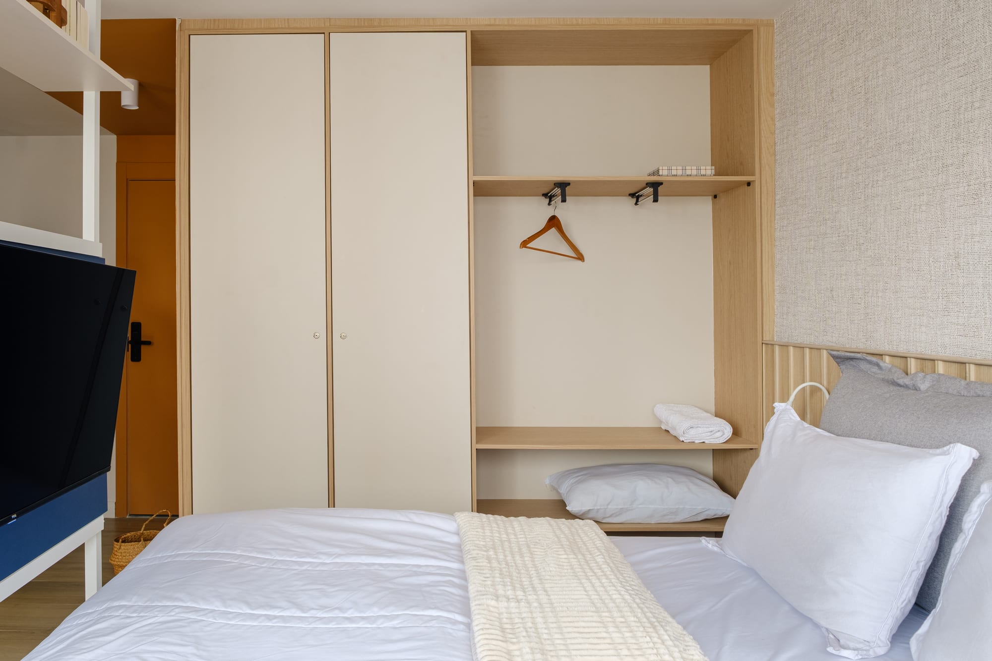 Bedroom with a white-dressed bed, light-wood wardrobe with an open niche and a TV on a blue panel.