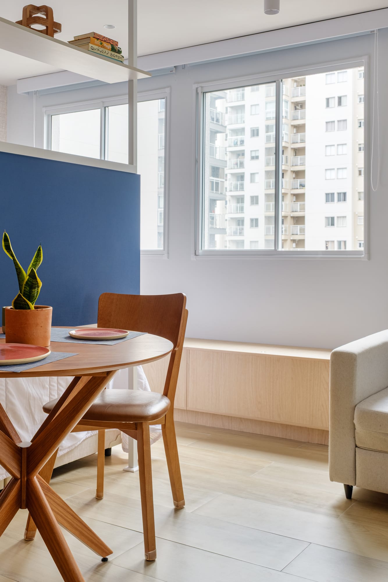 Dining area with a round wooden table, chairs, a light sofa and large windows overlooking the neighboring buildings.