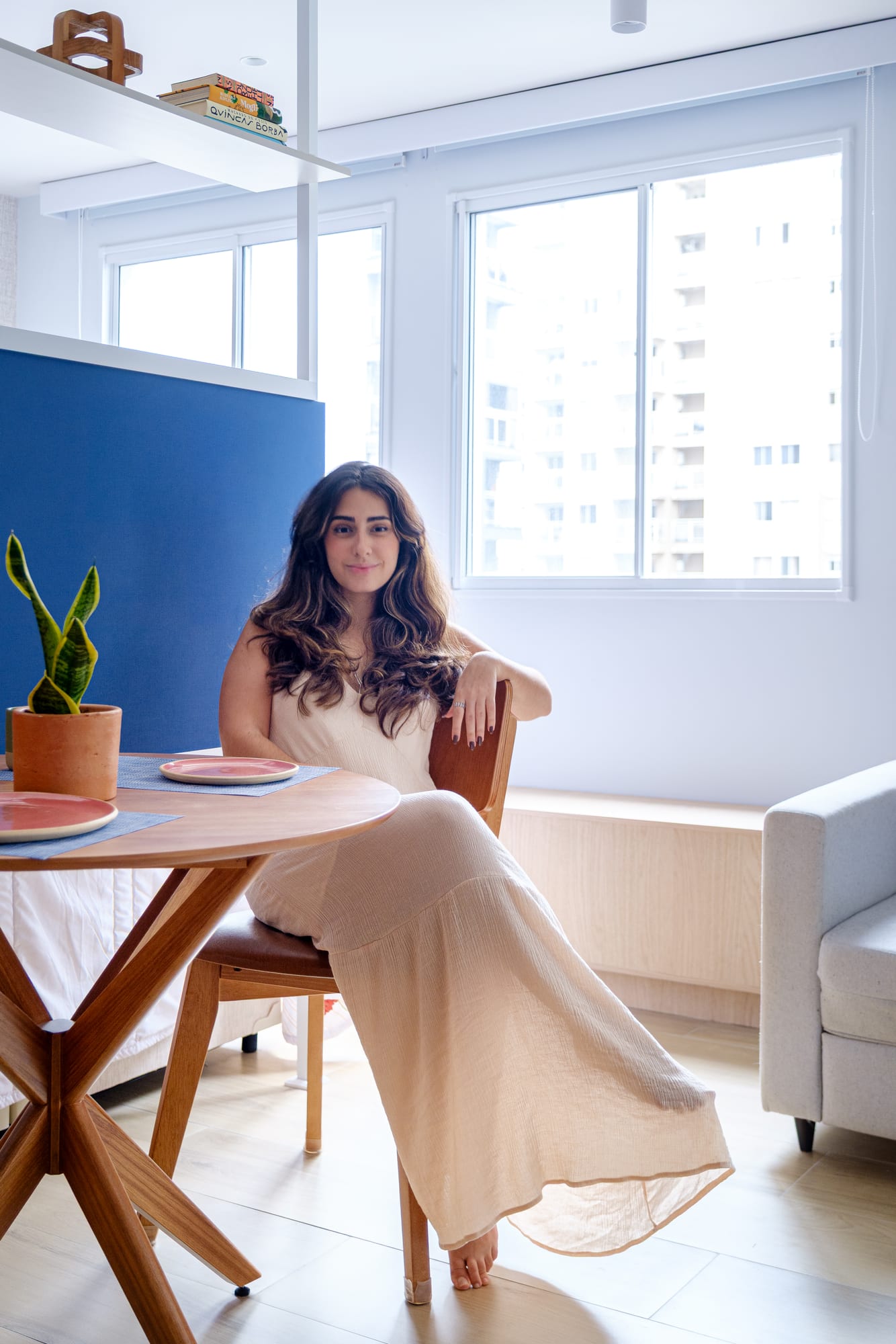 Woman in a light dress sitting at the round dining table, with the blue panel and the apartment windows behind her.