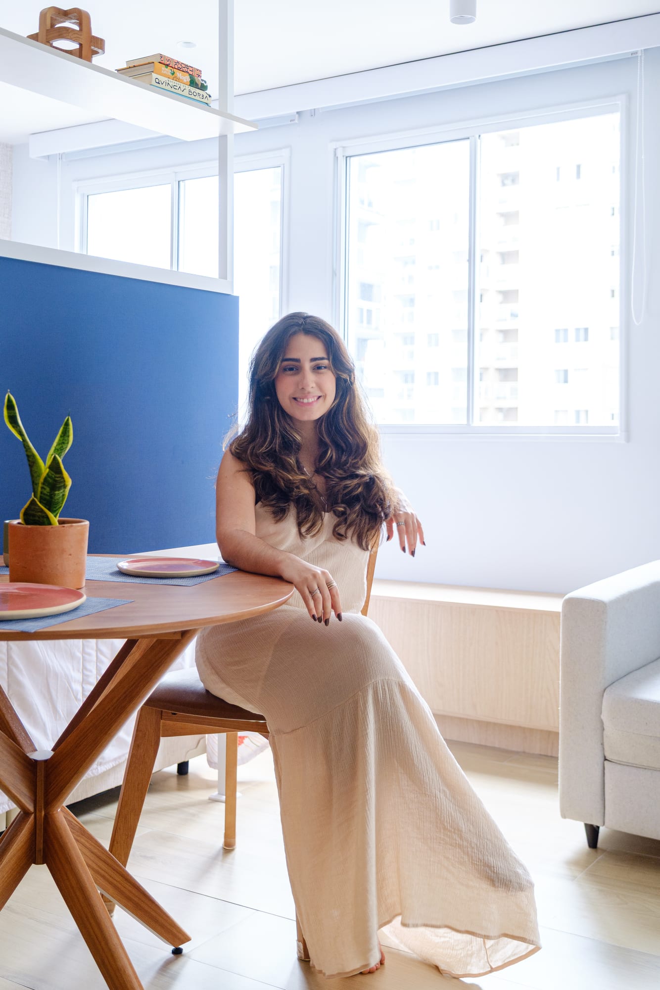 Portrait of a smiling woman at the dining table, with the sunlit living area and the light sofa beside her.