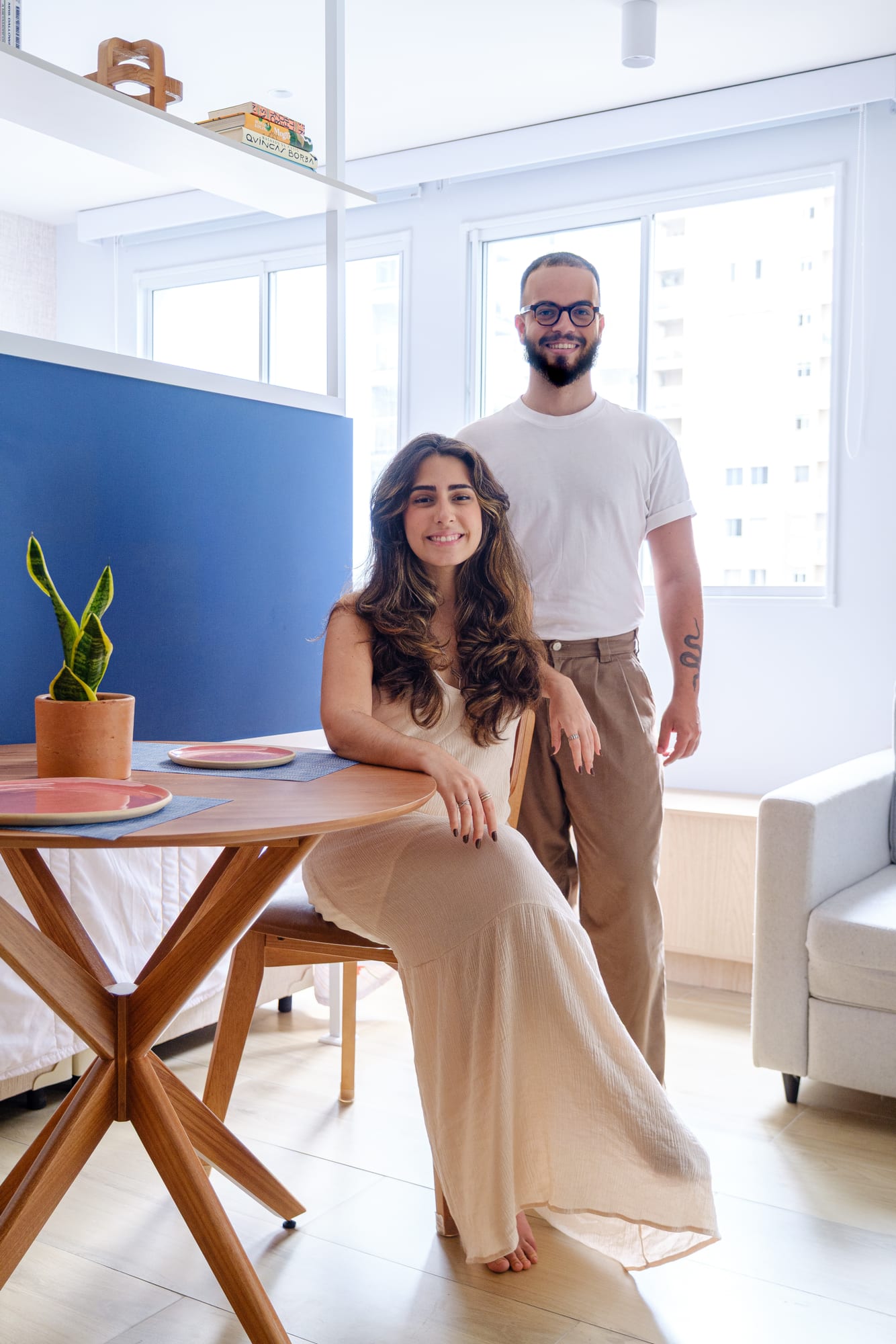 Couple posing in the apartment dining area, she seated at the round table and he standing beside her, the blue panel behind them.