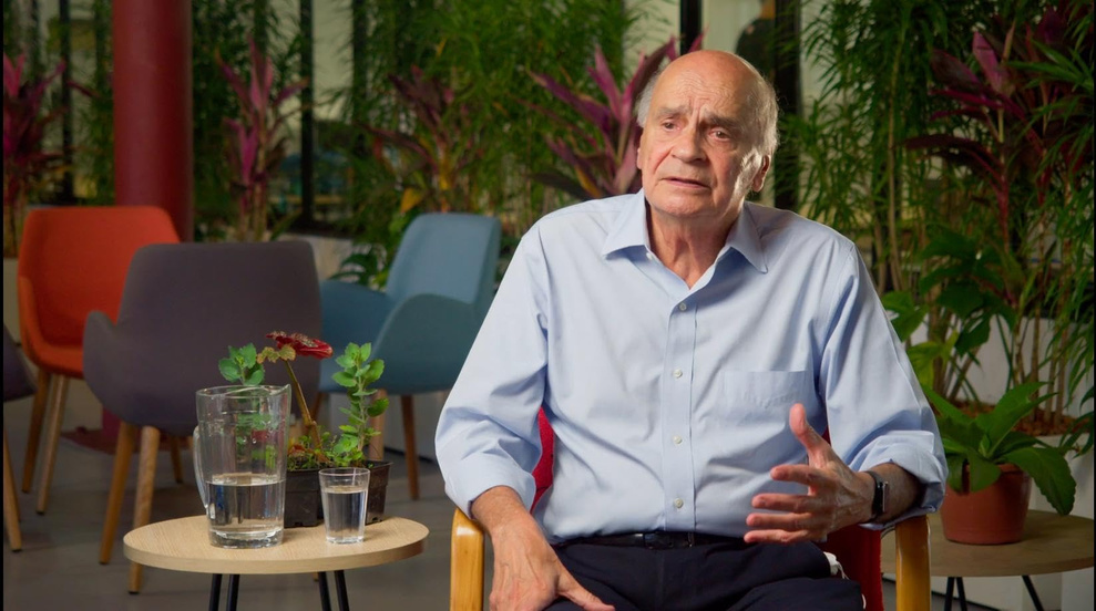 Homem idoso fala em um ambiente com plantas durante uma entrevista