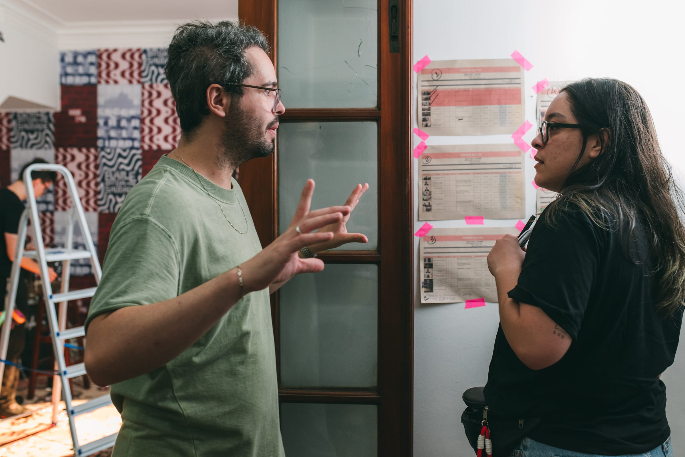 Homem de camiseta verde gesticula conversando com mulher de óculos e camiseta preta diante de cronogramas fixados com fita rosa.