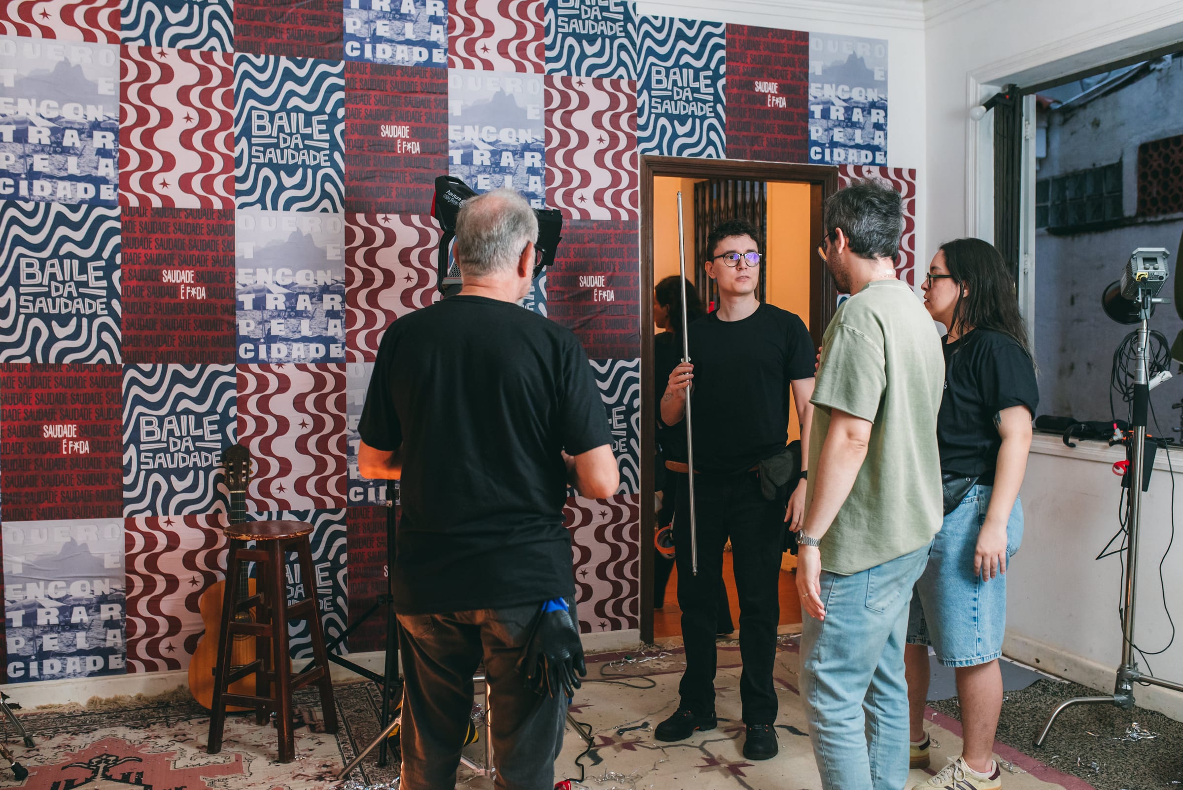 Quatro pessoas da equipe conversam em sala com painéis estampados Baile da Saudade, violão no banquinho e refletores.