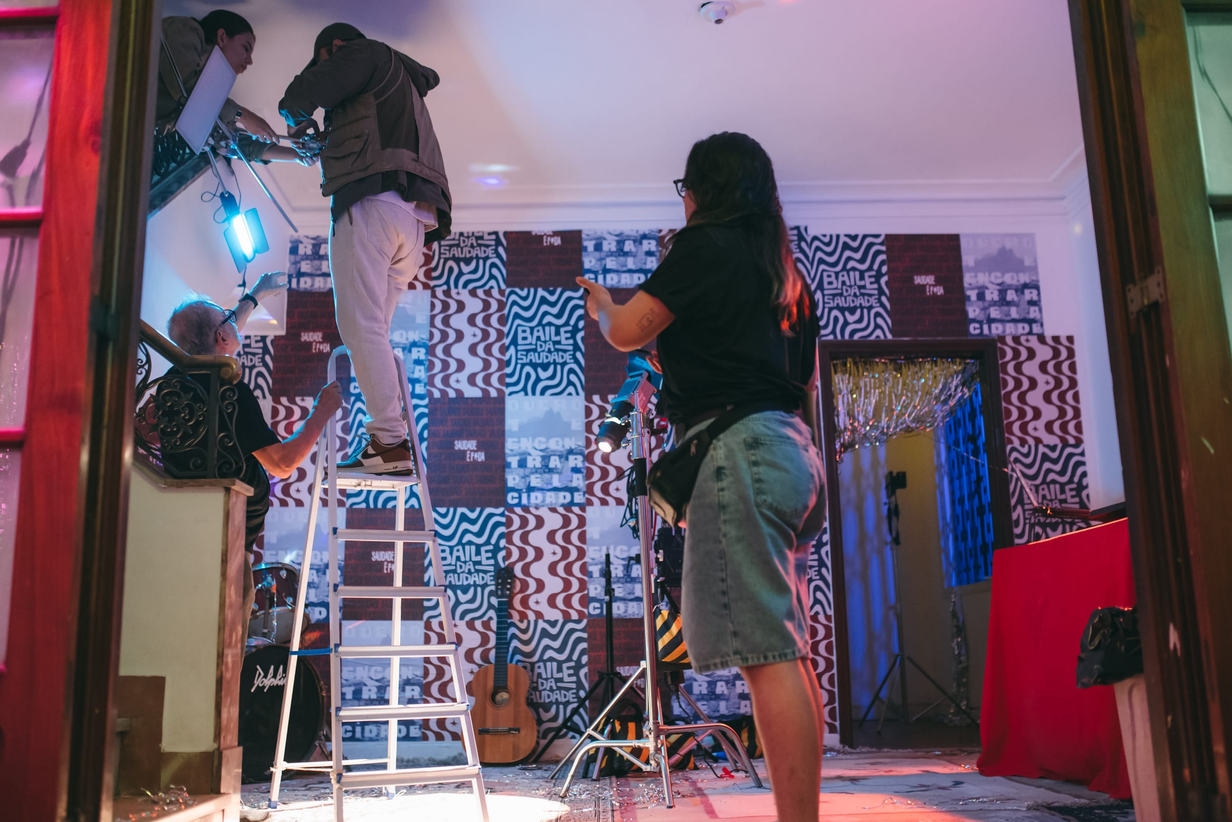Equipe monta iluminação com escada em sala de painéis estampados, com violão, bateria e mesa vermelha.