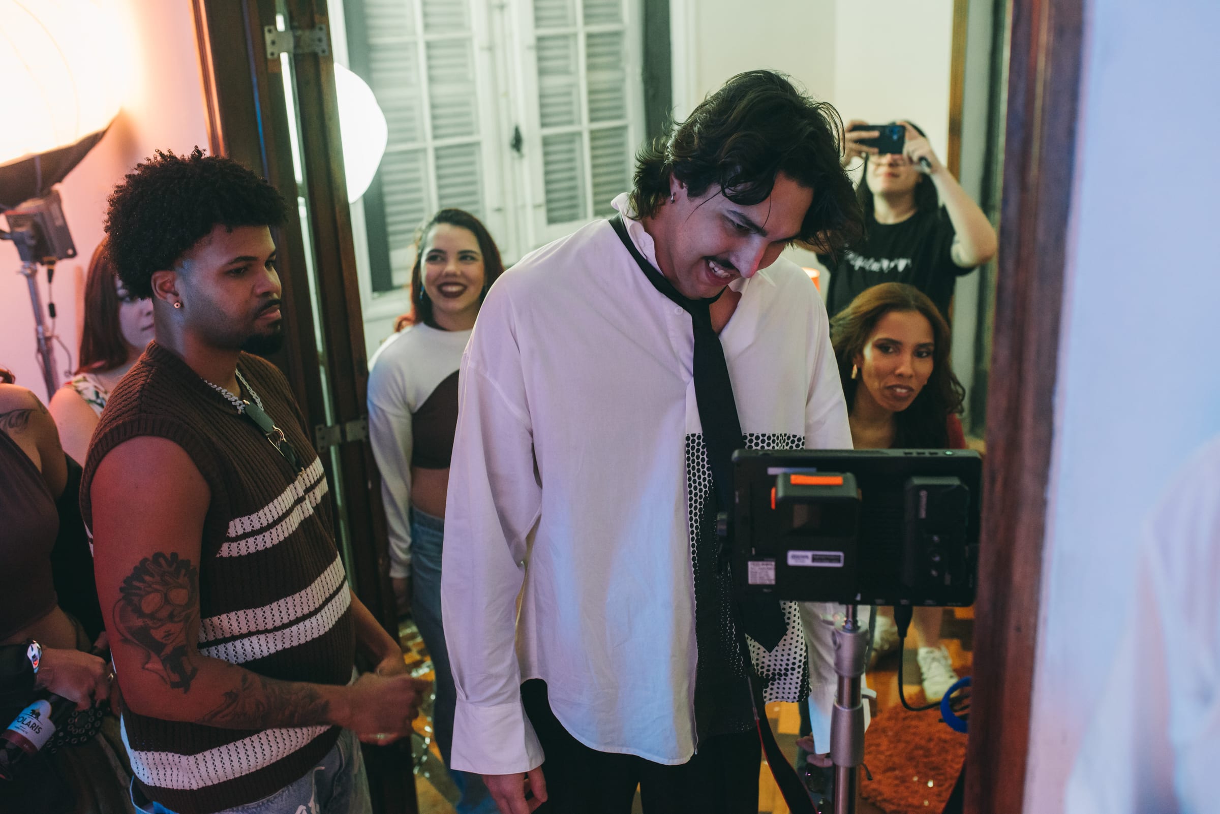 Cantor de camisa branca e gravata frouxa sorri diante do monitor de vídeo cercado pela equipe na porta iluminada.
