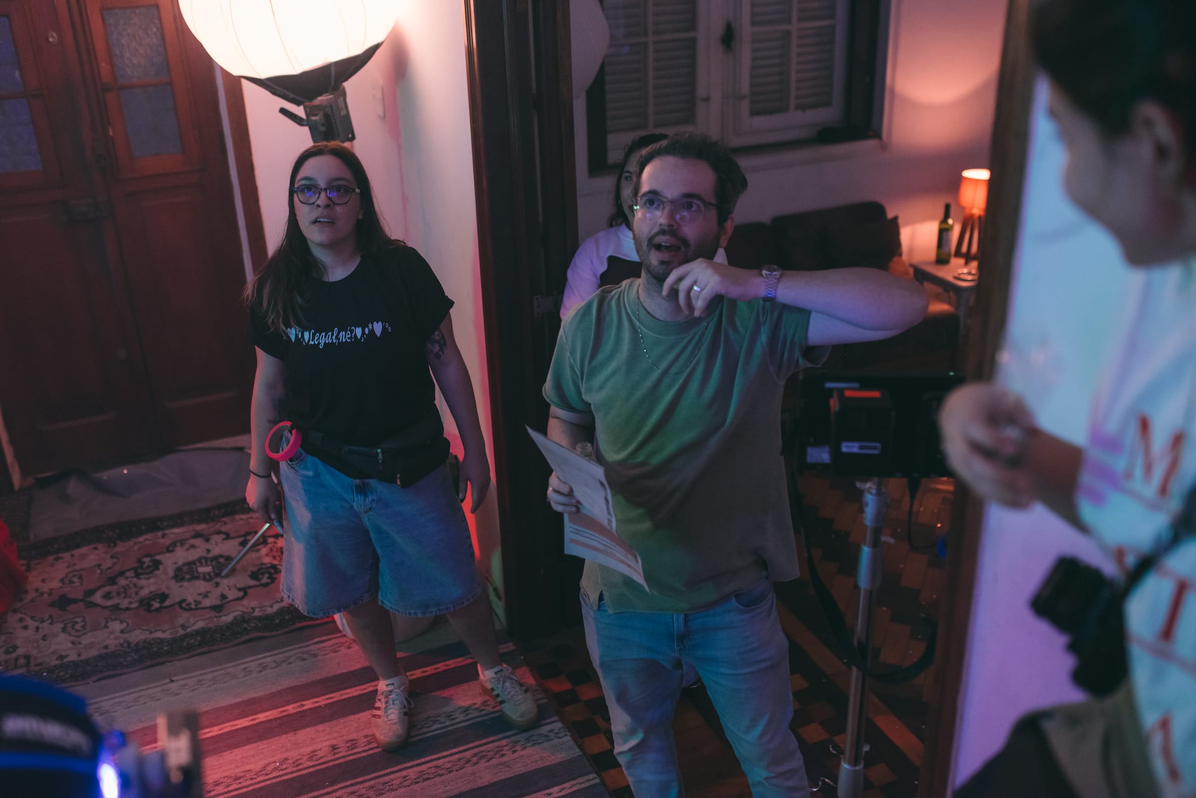 Diretor de camiseta verde segura roteiro e gesticula ao lado de assistente de camiseta preta sob luz rosada.
