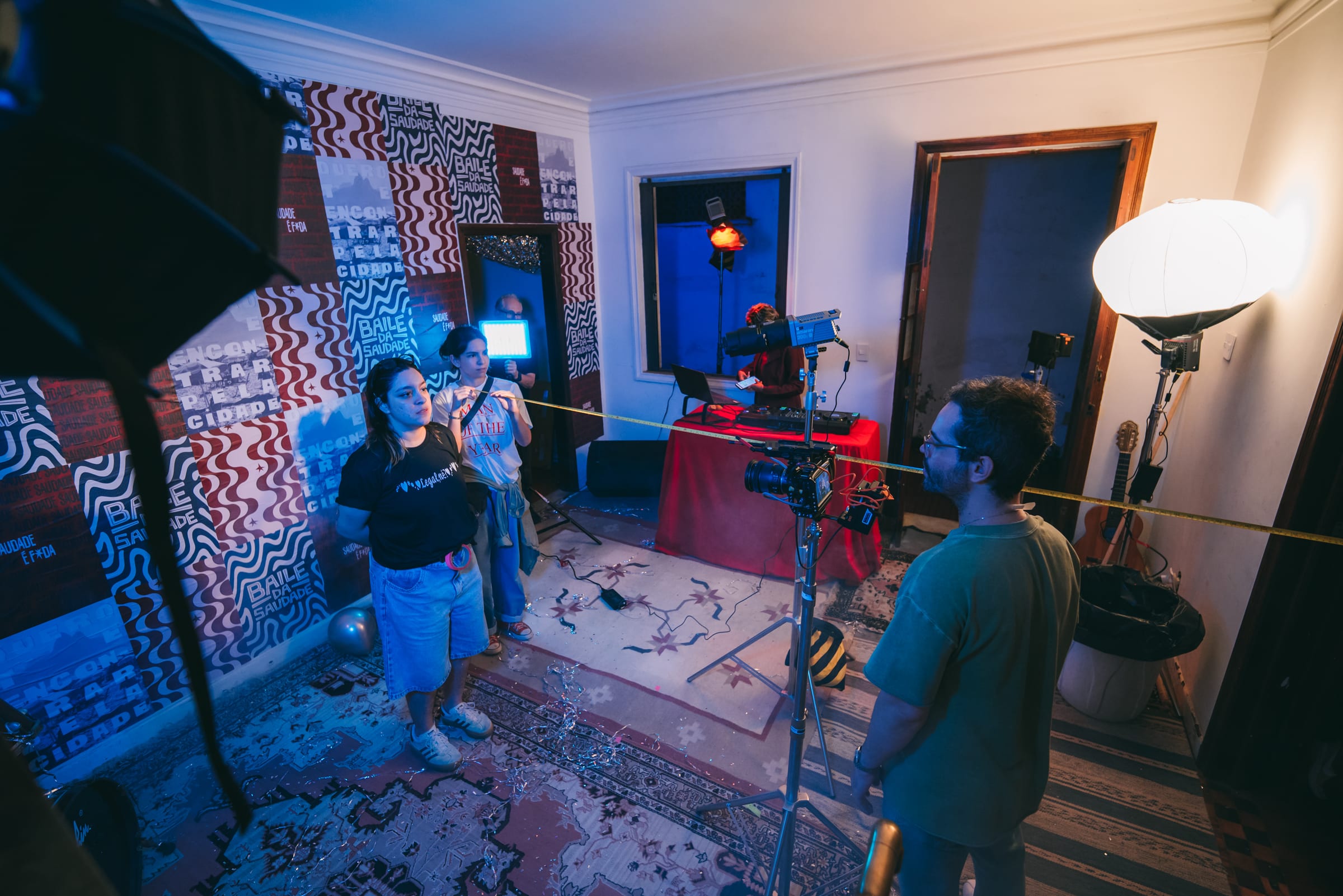Equipe estica trena em sala com mesa de DJ vermelha, câmera em tripé e painéis estampados sob luz azul.