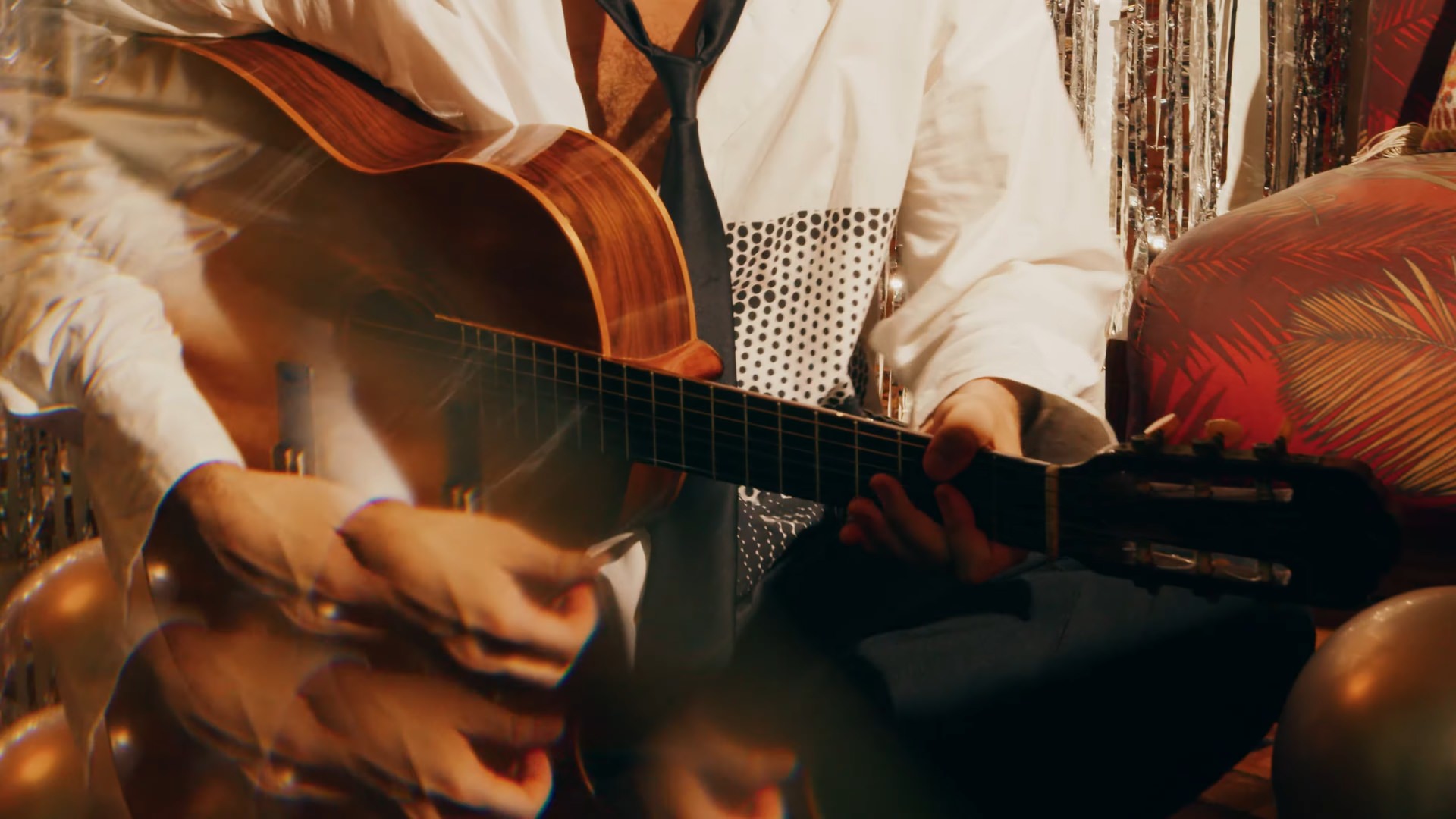 Mãos tocam violão acústico com músico de camisa branca e gravata, entre balões dourados e cortina prateada.