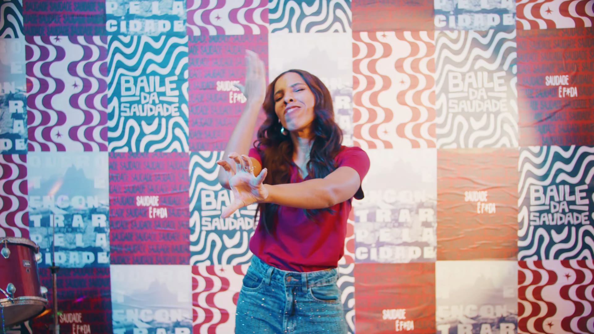 Mulher de camiseta vermelha e jeans dança de olhos fechados diante de parede de cartazes Baile da Saudade.