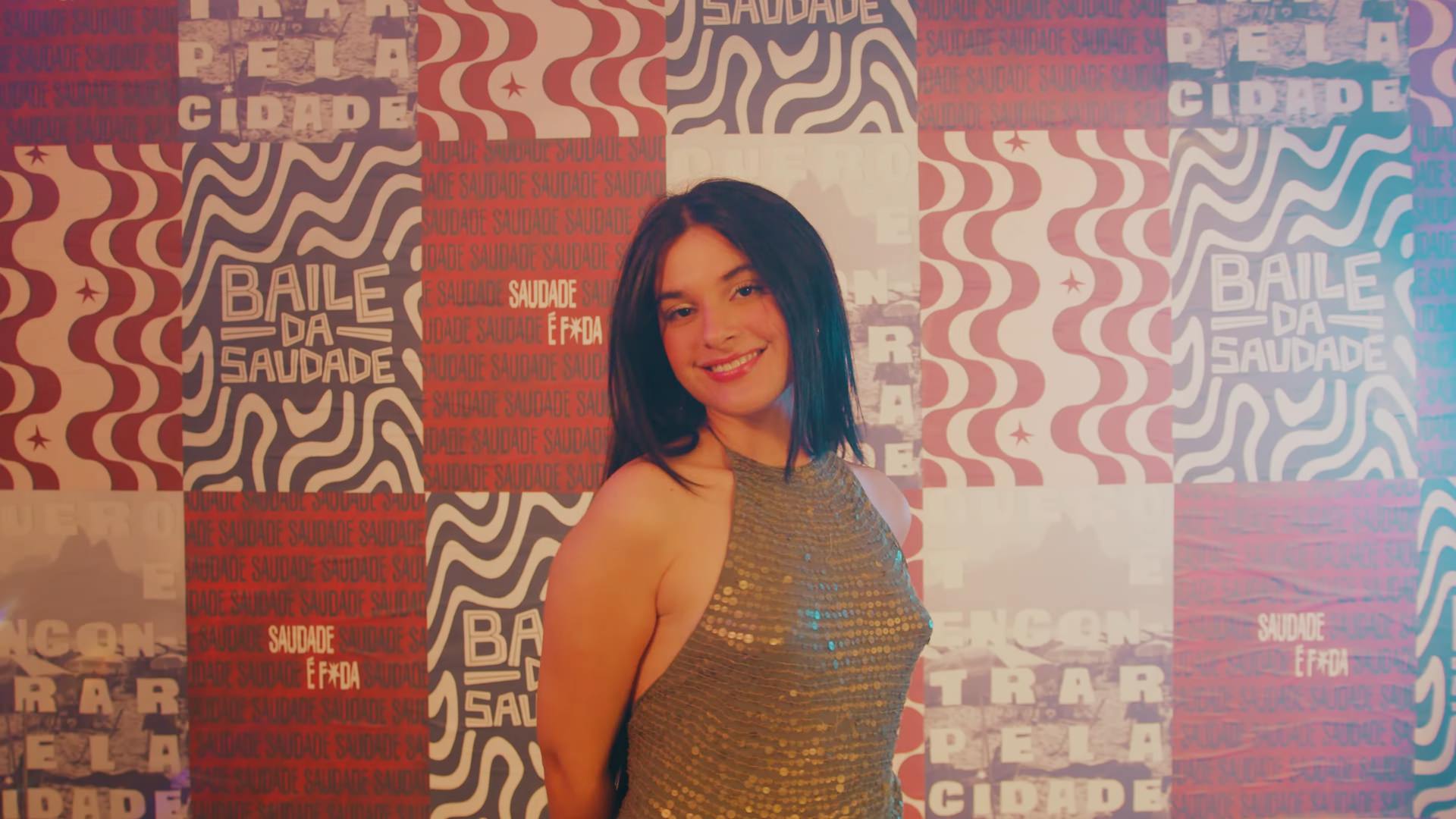 Mulher sorridente de blusa dourada de paetês posa diante de parede de cartazes Baile da Saudade.