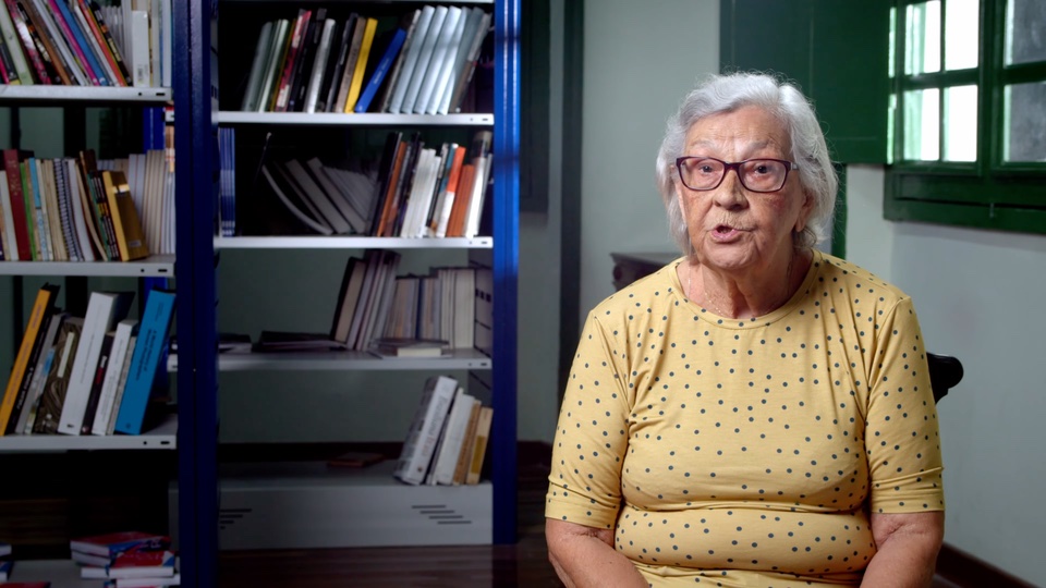 Mulher idosa fala diante de estantes de livros durante uma entrevista